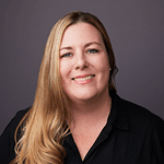 Professional headshot of a smiling woman with long blonde hair and a black top against a grey background.
