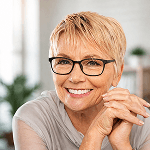 A smiling senior woman with short blonde hair and black glasses looks directly at the viewer, her hands clasped.