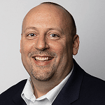 Smiling man with a bald head and goatee, wearing a dark suit jacket over a white shirt.
