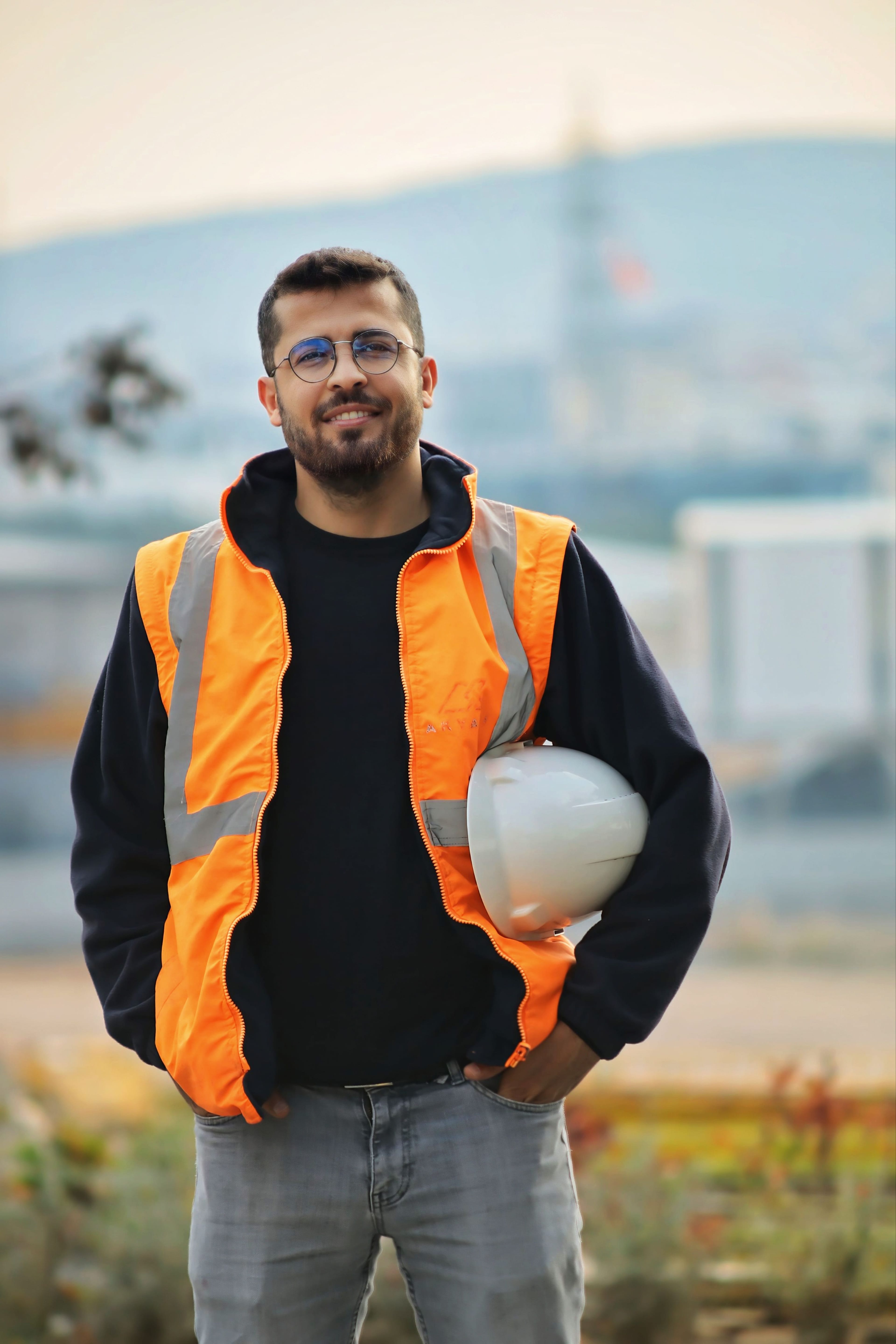 Smiling man in an orange safety vest and glasses holds a white hard hat, standing outdoors with a blurred industrial background.