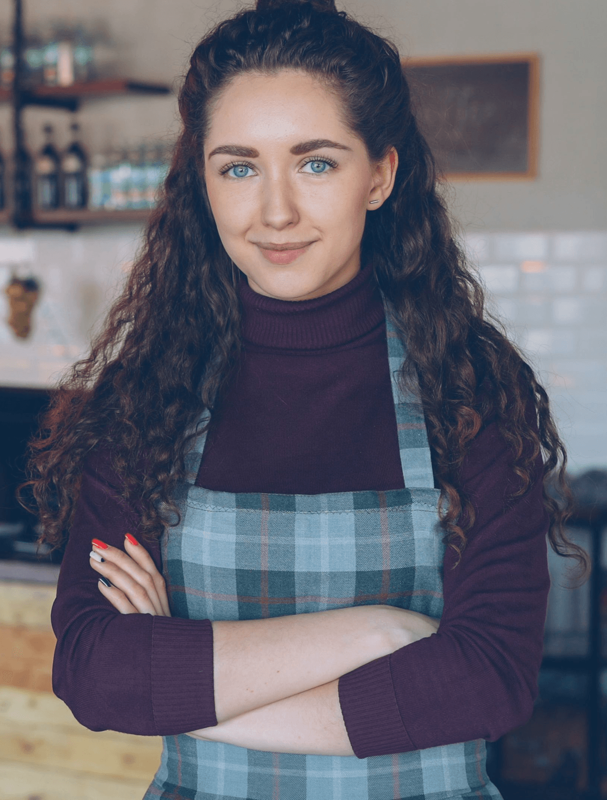 A smiling young woman with curly dark hair and blue eyes, wearing a dark red turtleneck and a plaid apron, stands with arms crossed in a blurry cafe setting.