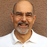 A cheerful bald man with round glasses and a gray goatee smiles directly at the camera, wearing a white polo shirt.