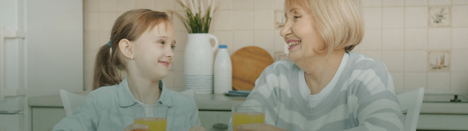 Smiling grandmother and granddaughter share orange juice at a kitchen table.
