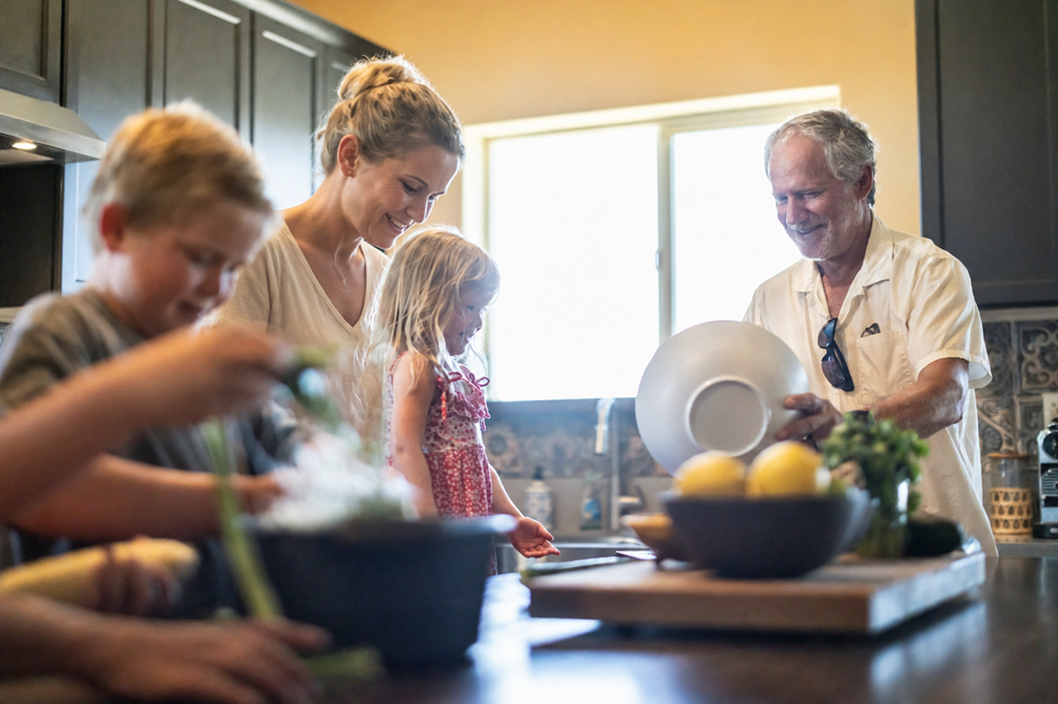 A family of four, including two adults and two children, happily preparing food together in a bright kitchen. A boy chops vegetables while a woman and girl smile, and an older man holds a bowl.