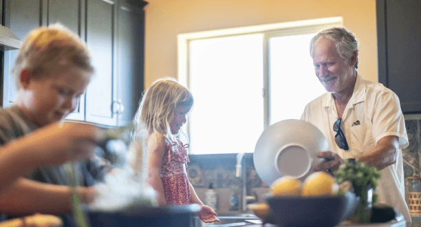 A smiling grandfather washes dishes in a sunlit kitchen while his young granddaughter watches and a boy works in the foreground, creating a warm, candid family atmosphere.