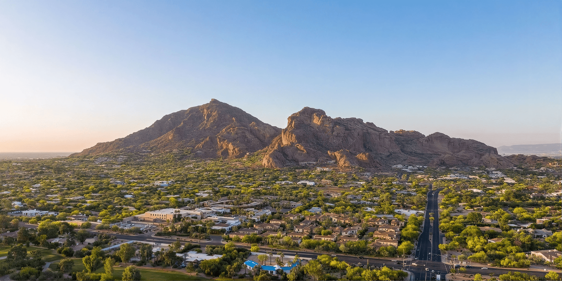 Aerial view of Camelback Mountain towering over a sprawling, tree-lined desert city in the soft light of sunrise or sunset.