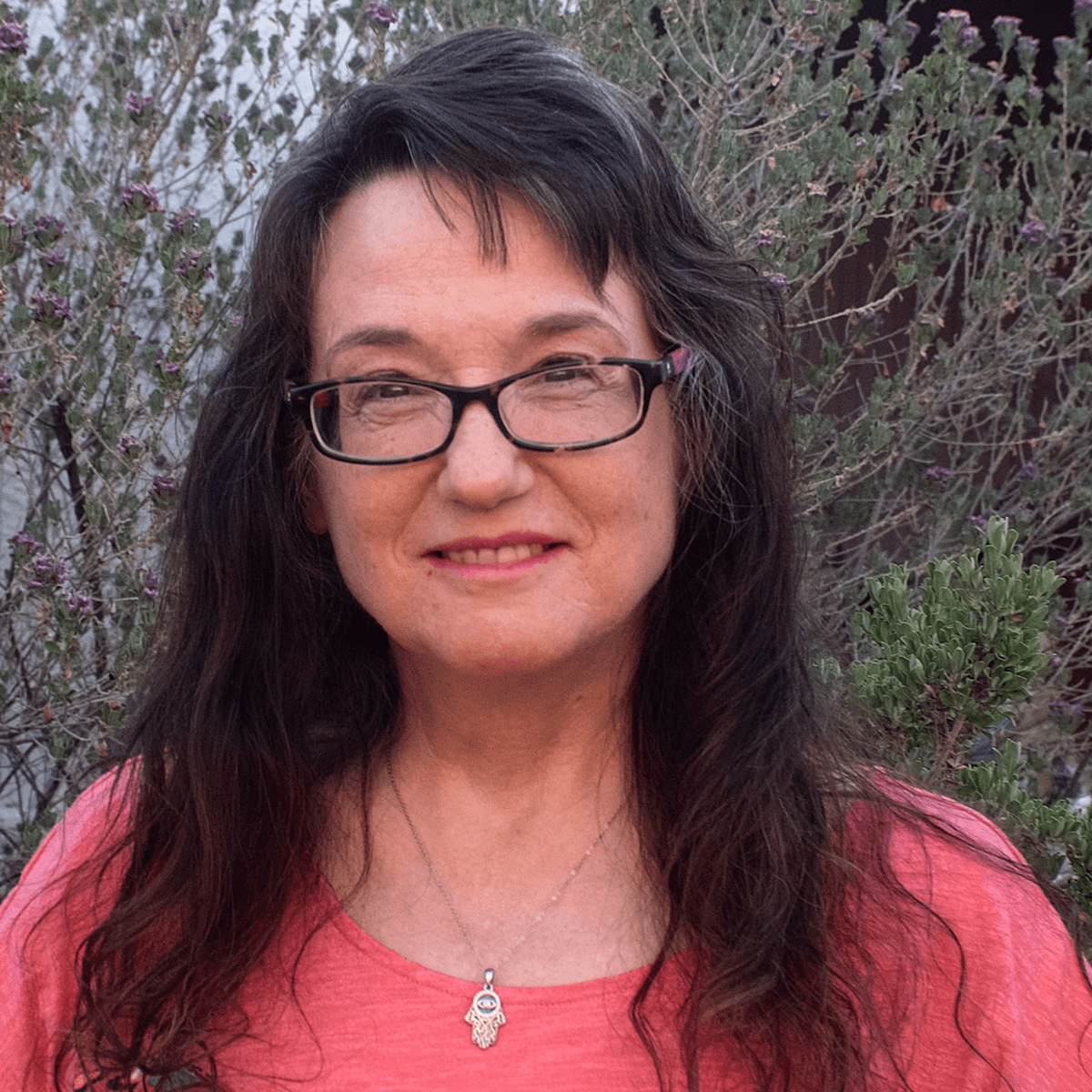 A smiling woman with long dark hair and glasses wears a peach top and hamsa necklace, against a blurred background of green and purple bushes.