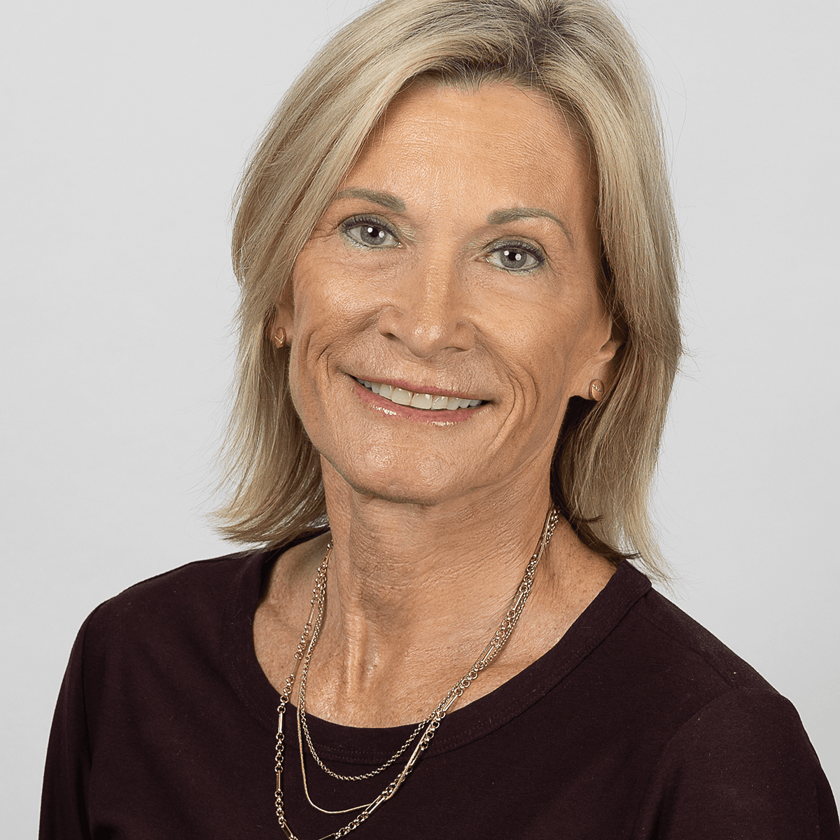 Smiling blonde woman with stud earrings and a layered gold necklace, wearing a dark top in a professional headshot.