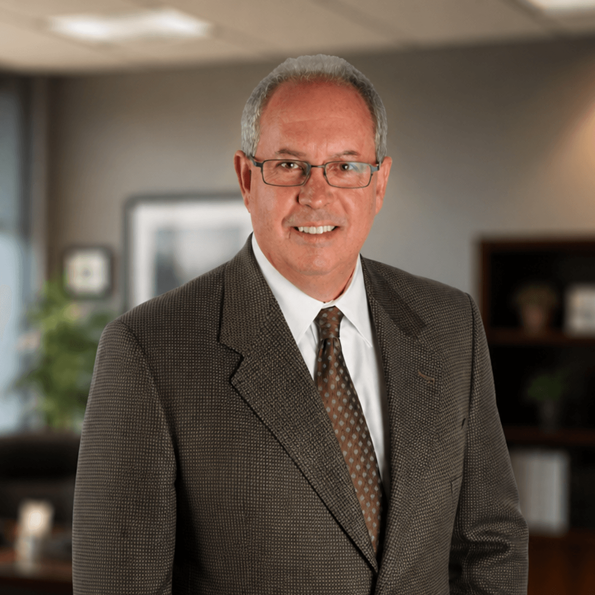 Smiling man in glasses and a brown suit with a patterned tie against a blurred office background.