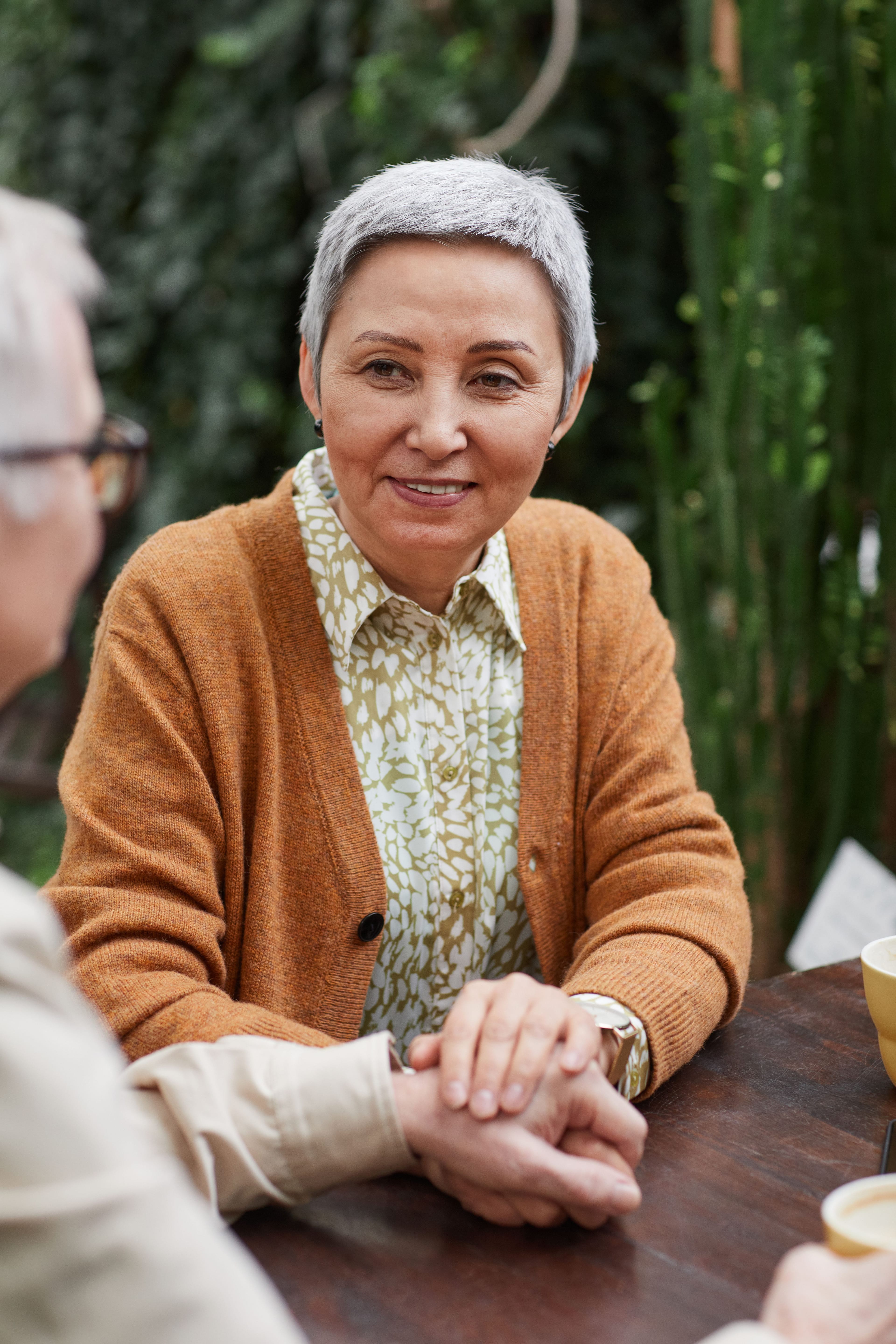 A smiling senior woman with short grey hair holds the hand of another person across a wooden table, with blurred green foliage in the background.