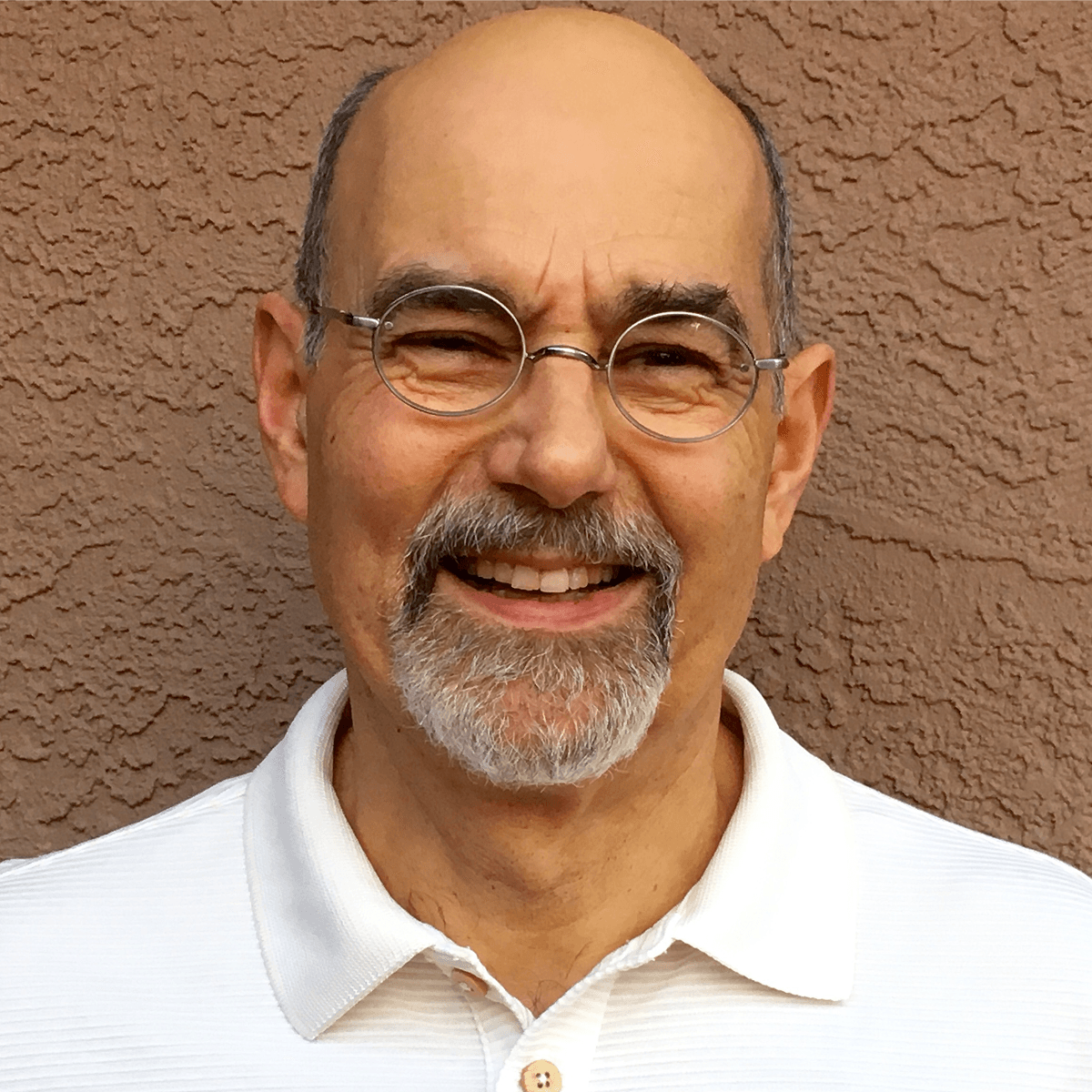 Smiling man with a bald head, grey beard and mustache, and round glasses wearing a white polo shirt against a textured brown wall.