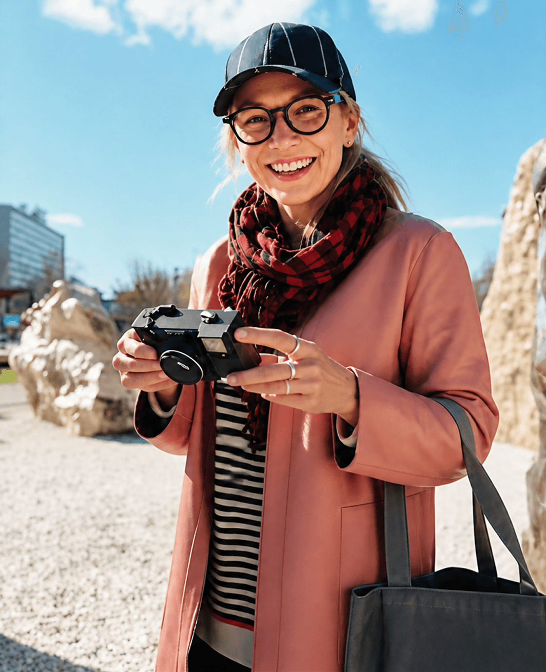 A smiling young woman in a striped cap, glasses, and pink coat holds a black film camera outdoors on a sunny day.